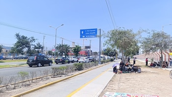 Personas sentadas a la sombra de los árboles en una acera junto a una avenida con tráfico de automóviles y un letrero azul