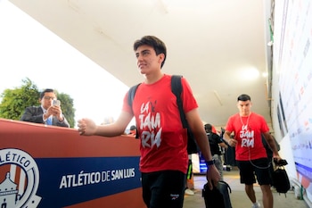 Joven con camiseta Reebok roja, mochila, camina en pasillo. Otro hombre con camiseta roja. Hombre de traje fotografía con móvil. Logo Atlético de San Luis