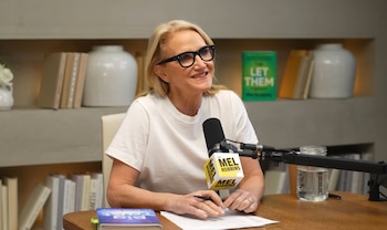Una mujer rubia con gafas y camiseta blanca sentada en una mesa de madera. Tiene un micrófono delante, papeles y estantes con libros y objetos detrás