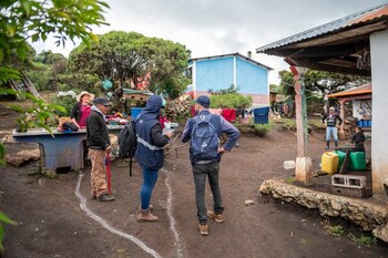 Grupo de personas en un entorno rural con casas de colores y vegetación. Dos individuos llevan chalecos y mochilas. Ropa tendida y botes de plástico visibles