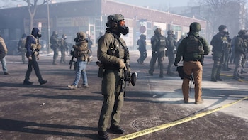 Varios agentes con equipo táctico completo y máscaras de gas en una calle urbana. Un oficial en primer plano usa una cámara corporal y sostiene un arma. Hay cinta policial en el suelo