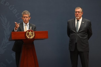 Hombre con gafas y traje oscuro habla en un podio de madera con emblema dorado; a su derecha, otro hombre con gafas y barba en traje