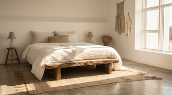 Dormitorio con cama de sábanas blancas, banco de madera rústica, alfombra de yute en suelo de hormigón pulido y una gran ventana que ilumina el espacio.
