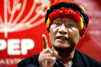 Primer plano de Alberto Pizango, un hombre indígena con una diadema tradicional roja, amarilla y negra, señalando con el dedo índice derecho. Fondo rojo borroso con logo blanco