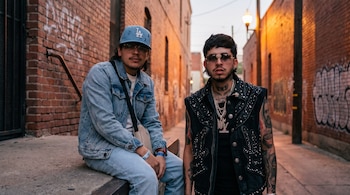 Dos hombres jóvenes con gafas en un callejón de ladrillo: uno viste chaqueta de mezclilla y gorra celeste, y el otro lleva un chaleco negro con tachas.