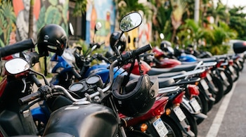 Fila de motocicletas estacionadas en una acera con manillares, espejos retrovisores y cascos. Al fondo se ven murales coloridos y vegetación.