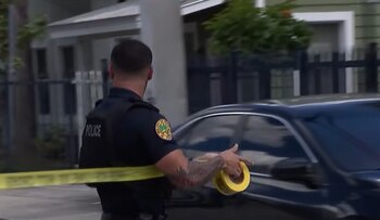 Un agente de policía visto de espaldas coloca cinta amarilla de precaución frente a un automóvil negro y una casa