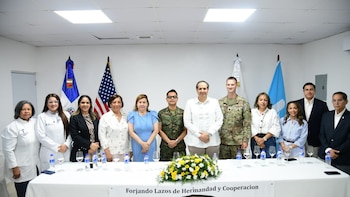 Catorce personas, entre personal médico y militar, posan detrás de una mesa con arreglo floral, botellas de agua y copas. Banderas de dos países al fondo