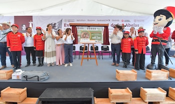 Grupo de personas en un escenario, incluyendo niños con uniforme rojo y un personaje de mascota, aplaudiendo junto a un billete gigante de la Lotería Nacional