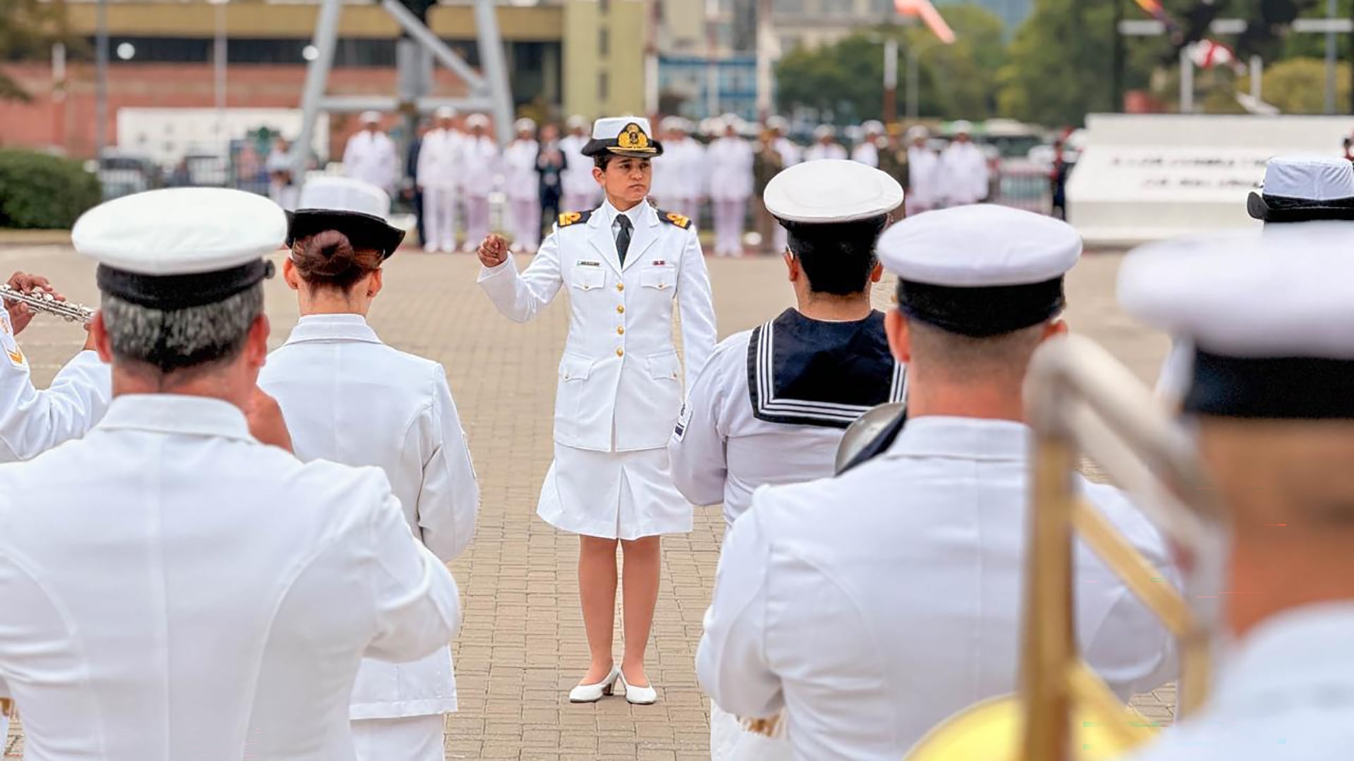 Una oficial naval dirige a la banda militar durante una formación al aire libre, con los músicos en primer plano tocando sus instrumentos, en el homenaje a Guillermo Brown
