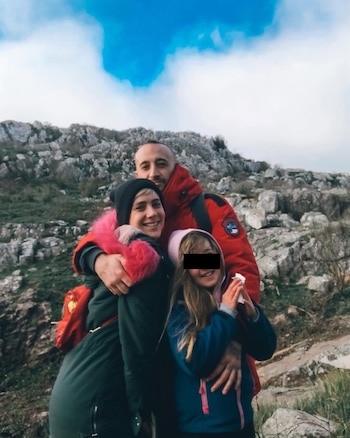 Una mujer, un hombre y una niña posan abrazados en un paisaje exterior con montañas rocosas y cielo nublado. La mujer lleva un gorro negro con pelo rosa y el hombre una chaqueta roja