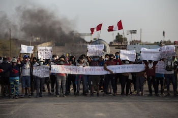 Multitud de personas con pancartas y una bandera, algunas con mascarillas, bloqueando una vía. Dos banderas peruanas y humo oscuro son visibles en el fondo