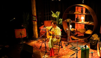 Un hombre toca guitarra acústica sentado en un escenario oscuro con un micrófono, un teclado y un estante circular decorado con objetos y una planta