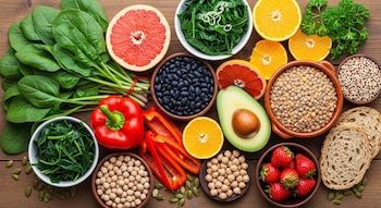 Vista aérea de espinacas, pimiento, cítricos, aguacate, frijoles, lentejas, garbanzos, quinua, fresas, pan y semillas de calabaza sobre madera.