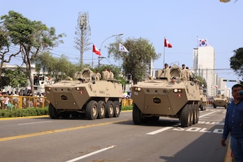 Dos vehículos blindados de color beige con soldados, varias banderas de Perú y una de Corea del Sur, multitud, edificios, árboles, calle con señalización "SOLO BUS"