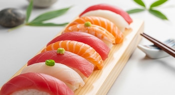 Imagen de varios nigiris de sushi alineados sobre una bandeja de madera, con arroz brillante y láminas de atún, salmón, gamba y pescado blanco. Hay palillos al fondo.