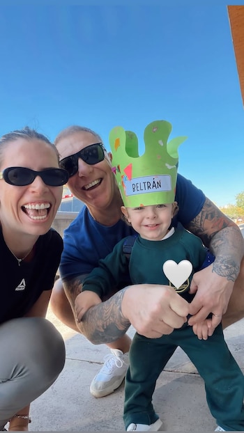 Fotografía de una familia feliz al aire libre: una mujer con gafas, un hombre tatuado con gafas y un niño sonriente con una corona verde que dice Beltrán