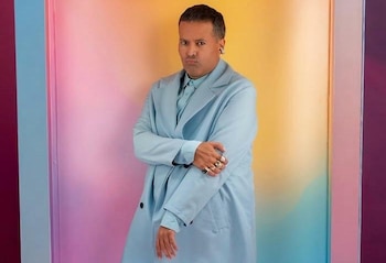 Hombre de piel morena con traje celeste, camisa y corbata. Brazos cruzados. Anillos y pendientes en un fondo de colores degradados
