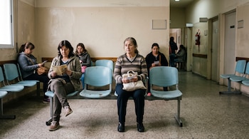 Mujeres sentadas en sillas plásticas azules dentro de una sala de espera con paredes claras y un pasillo al fondo.