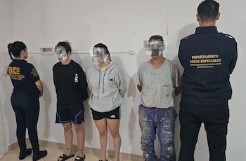 Dos oficiales de policía, una de frente y uno de espaldas, junto a tres personas con rostros borrosos, dos mujeres y un hombre, contra una pared blanca