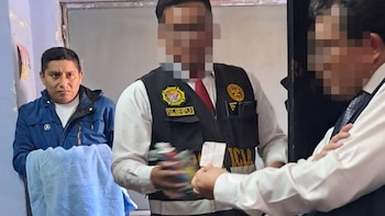 Un hombre con chaqueta azul permanece de pie junto a policías con rostros difuminados que sostienen paquetes incautados