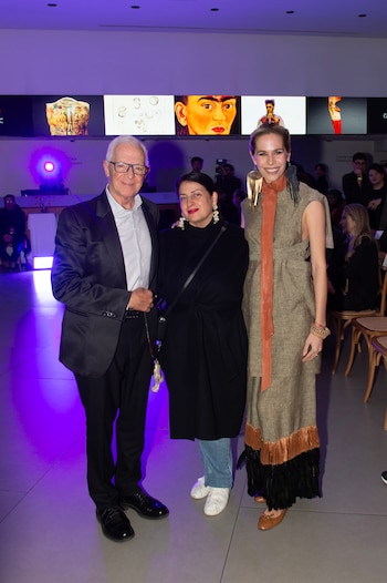 Un hombre con traje y dos mujeres con ropa elegante posan de pie frente a una pantalla iluminada con retratos