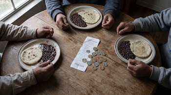 Vista cenital de una mesa de madera con tres platos de frijoles y tortillas, manos de adultos apoyadas, un recibo de supermercado arrugado y varias monedas dispersas.