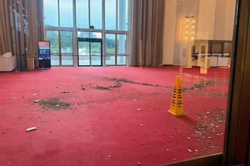 Escombros y tierra están esparcidos sobre una alfombra roja junto a un cono amarillo de advertencia en un vestíbulo con amplios ventanales
