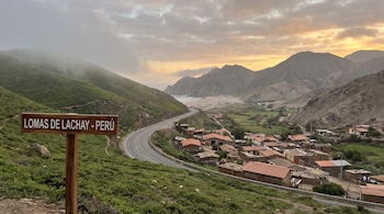 Cartel de madera con texto "LOMAS DE LACHAY - PERÚ", carretera, colinas verdes cubiertas de vegetación, un pueblo con casas de techos naranjas, montañas, océano, cielo nublado.