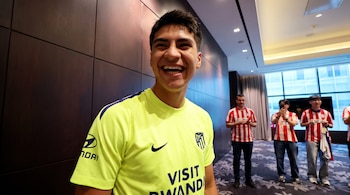 Un joven sonríe vistiendo una camiseta de fútbol amarilla con logos. Detrás, tres hombres con camisetas de rayas rojas y blancas. Pared de madera al fondo.