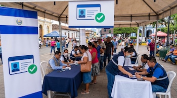 Vista de una jornada de renovación de DUI en El Salvador, con mesas de atención, personal y ciudadanos haciendo fila bajo carpas. Una bandera de El Salvador es visible.