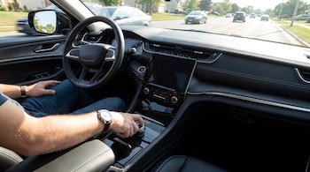 Interior de un vehículo con un conductor manejando, mostrando el volante, el tablero y la mano del conductor sobre el selector giratorio de cambios.
