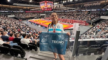 Una joven sonriente sostiene en las tribunas de un estadio una bandera azul con los nombres de las marcas Cochlear y Tecno salud.
