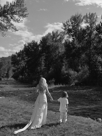 Fotografía en blanco y negro de mujer y niño de espaldas caminando tomados de la mano junto a un río, con árboles en el fondo y cielo nublado