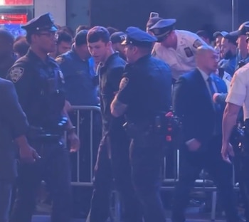 Un joven con camiseta oscura es escoltado por dos agentes de policía uniformados entre una multitud y barreras metálicas
