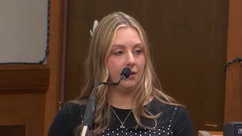 Una mujer rubia vestida de negro habla ante un micrófono frente a un fondo de madera