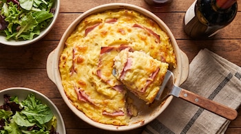 Pastel de papas con jamón y queso gratinado en fuente, porción siendo servida con espátula, dos cuencos de ensalada, botella de vino tinto y servilleta sobre mesa de madera.