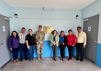 Ocho personas, entre ellas siete civiles y un militar uniformado, posan alineadas frente a una pared con una placa dorada entre dos puertas