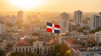 Bandera de República Dominicana con escudo ondeando en asta sobre un edificio colonial. Al fondo, rascacielos y ciudad bajo luz dorada de amanecer o atardecer.