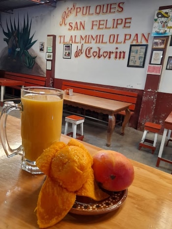 Una jarra de vidrio con bebida amarilla, un mango cortado y otro entero en un plato de barro sobre una mesa de madera dentro de una estancia