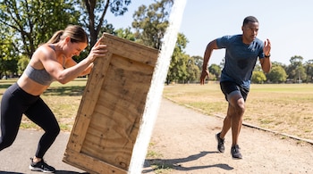 Una mujer vestida con ropa deportiva empuja una estructura de madera y un hombre corre por un camino en un parque.