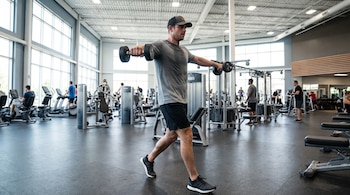 Un hombre con gorra y ropa deportiva sostiene dos mancuernas con los brazos extendidos hacia los lados en un gimnasio con máquinas.