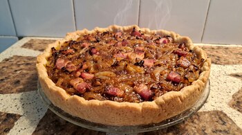 Tarta circular horneada con relleno de cebolla dorada y trozos de panceta sobre un plato transparente de vidrio.