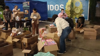 Varias personas organizan donaciones en cajas de cartón. Se observan productos diversos, mesas, carpas azules y carteles en un espacio exterior
