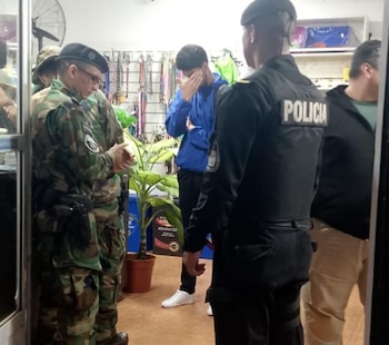 Tres hombres dentro de un local: uno con uniforme de camuflaje, otro con uniforme de policía de espaldas, y un civil de azul cubriéndose la cara