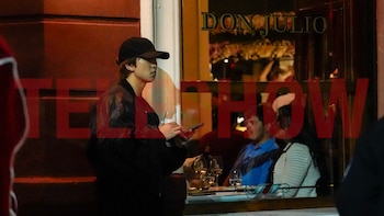 Un hombre con gorra negra y copa en mano frente a la ventana del restaurante Don Julio, mientras comensales cenan en el interior