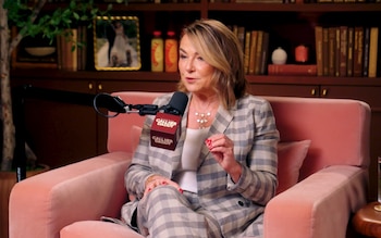 Mujer de cabello rubio, vestida con un traje a cuadros gris y blanco, sentada en un sillón rosa, habla en un micrófono con el logo de Call Her Daddy