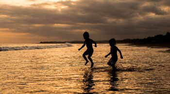 Siluetas de dos niños saltando y chapoteando en la orilla del mar al atardecer, con el agua reflejando tonos dorados y el cielo nublado.