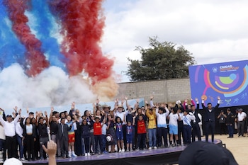Personas sobre un escenario levantan las manos ante columnas de humo rojo, azul y blanco junto a un cartel de la Ciudad Deportiva