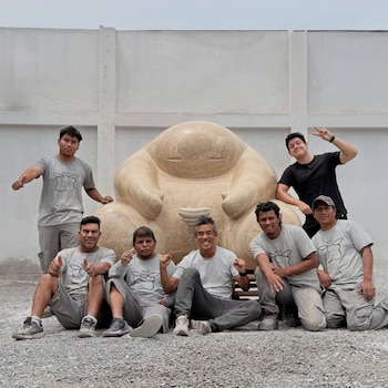 Siete hombres posan junto a una escultura beige de forma redondeada ante una pared blanca sobre un terreno de gravilla
